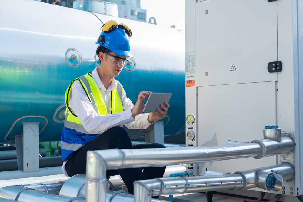 Technician wearing a hard hat and safety gear inspecting industrial pipework and tanks using a tablet, ensuring compliance standards for water hygiene service